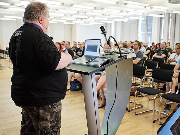 Frank Berger steht an einem Rednerpult mit Laptop und Mikrofon und hält einen Vortrag vor einem Publikum in einem modernen, hellen Seminarraum. Das Publikum hört aufmerksam zu. Der Vortrag behandelt das Thema „Running AI Models Inside PHP“ und die Integration von KI in TYPO3.